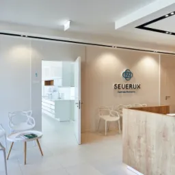 Jasne, minimalistyczne wnętrze centrum medycznego z recepcją z drewnopodobnym wykończeniem, białymi krzesłami w poczekalni i logo firmy na ścianie.