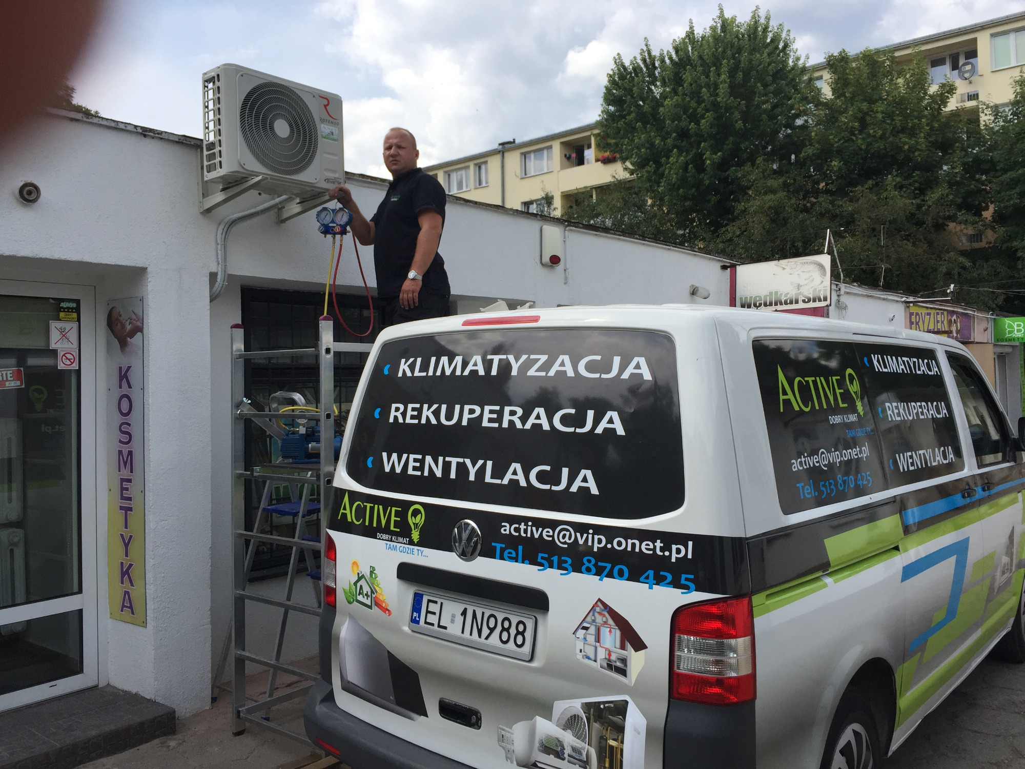 Mężczyzna na drabinie serwisuje zewnętrzną jednostkę klimatyzacji Rotenso przy budynku, na pierwszym planie samochód firmowy z reklamą klimatyzacji, rekuperacji i wentylacji.