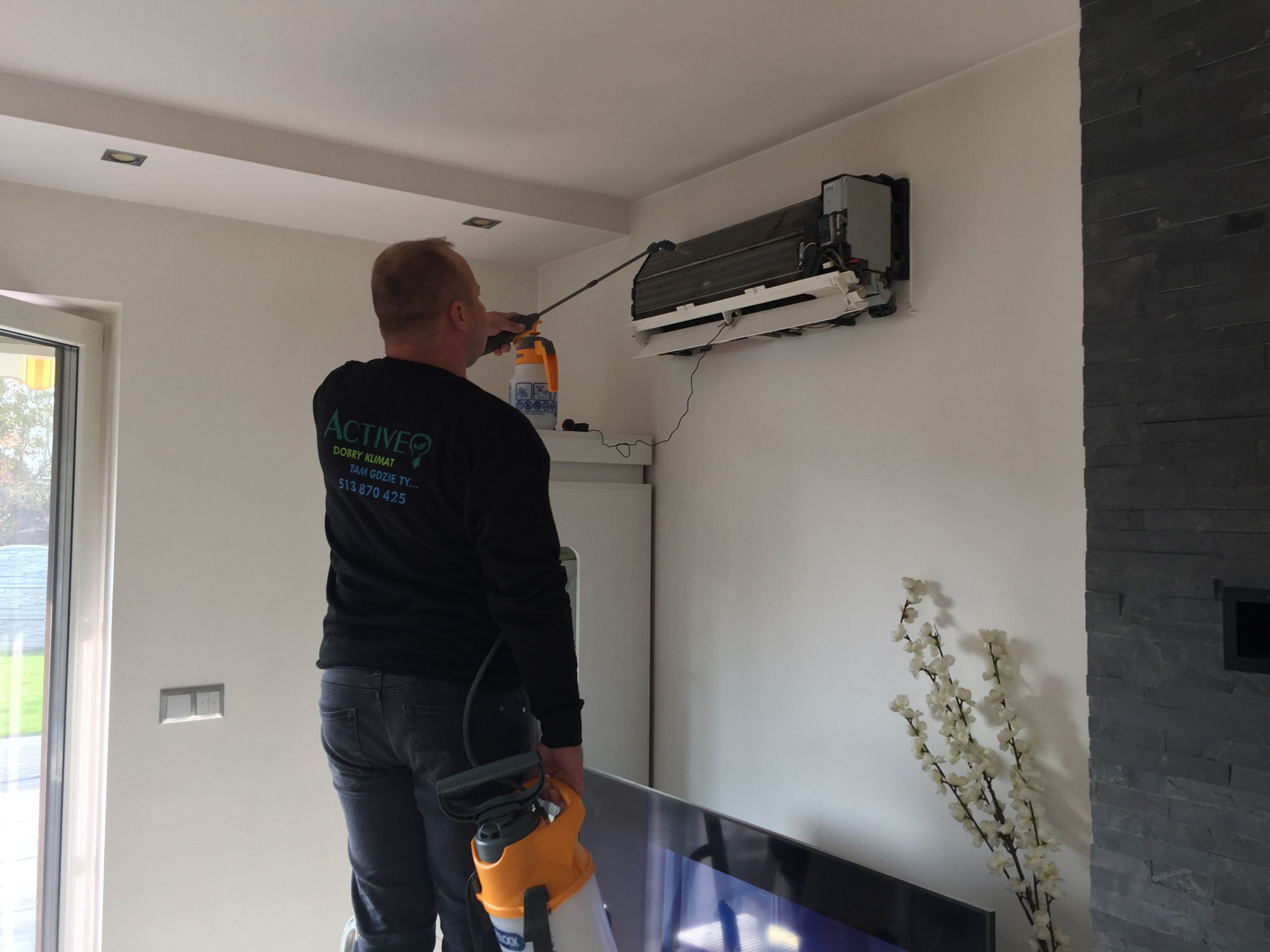 Mężczyzna w czarnej bluzie z logo firmy spryskuje wnętrze klimatyzatora zawieszonego na ścianie w nowoczesnym salonie, używając spryskiwacza ciśnieniowego z długą lancą.
