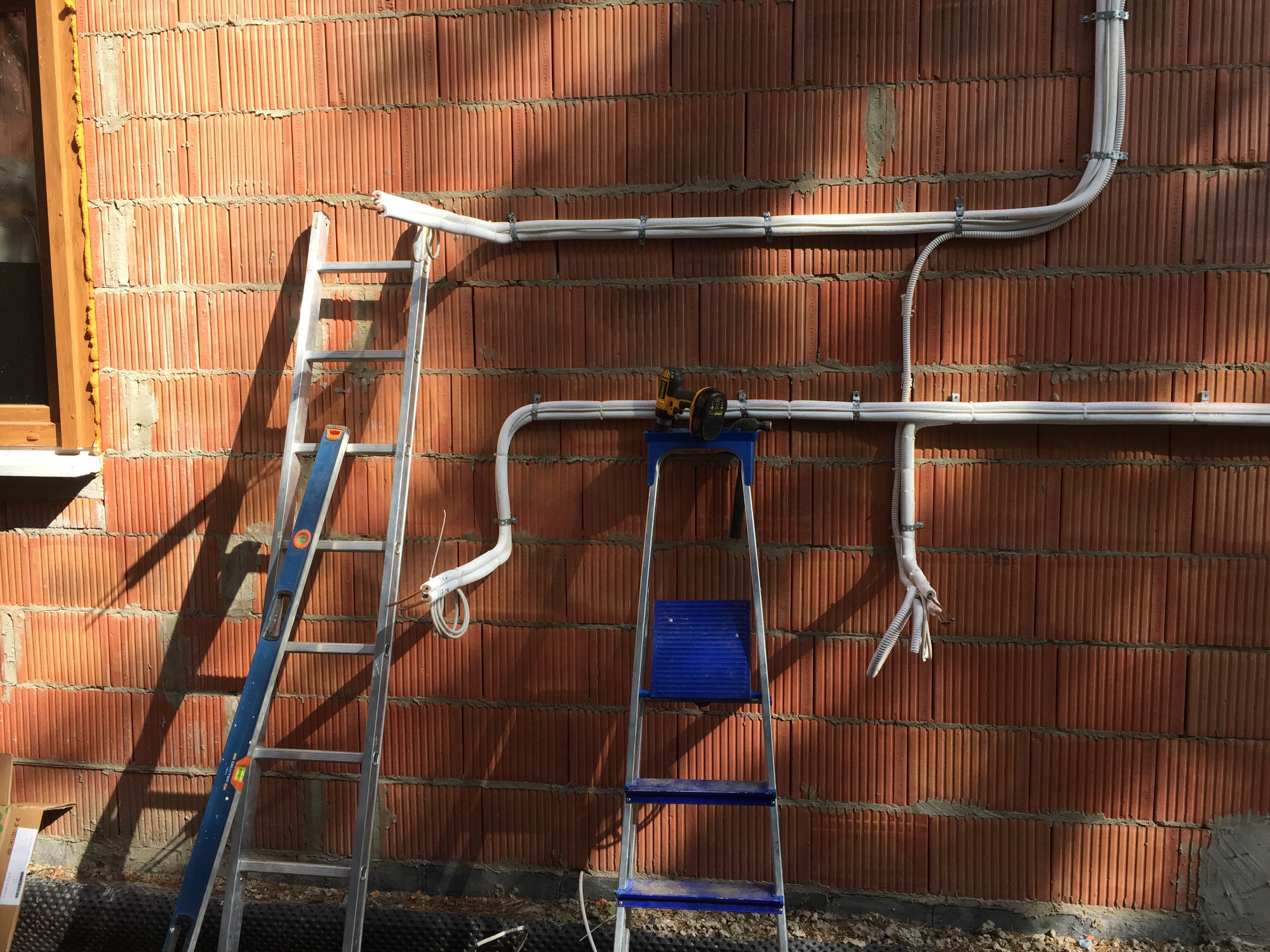 Montaż białych rurek instalacyjnych na ceglanej ścianie, widoczne drabiny i narzędzie na stołku.