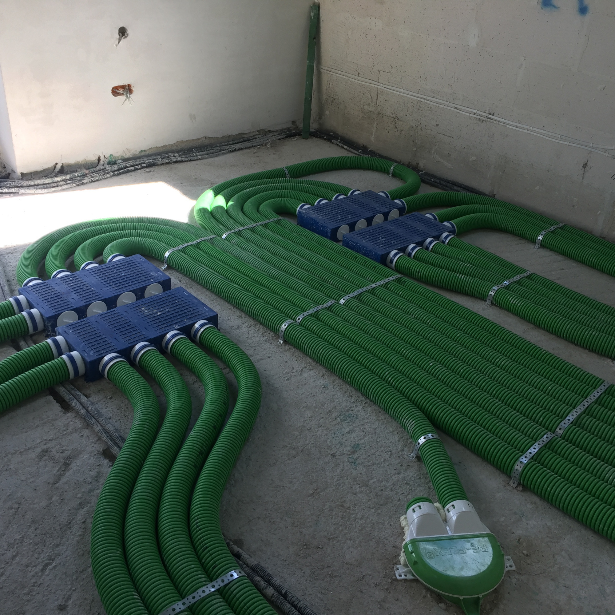 Rozprowadzenie zielonych rur wentylacyjnych w surowym stanie budynku, widoczne niebieskie skrzynki rozdzielcze i mocowania metalowe.