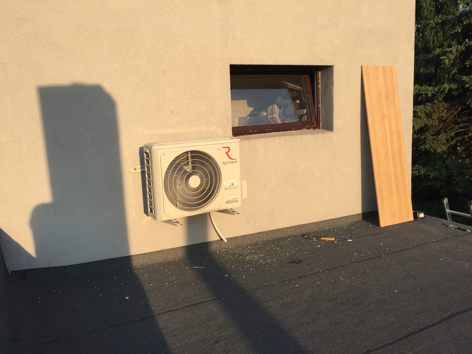 Zewnętrzna jednostka klimatyzacji Rotenso zamontowana na elewacji budynku, widoczna mała okiennica, deska oparta o ścianę, narzędzia i białe opiłki na czarnym dachu.