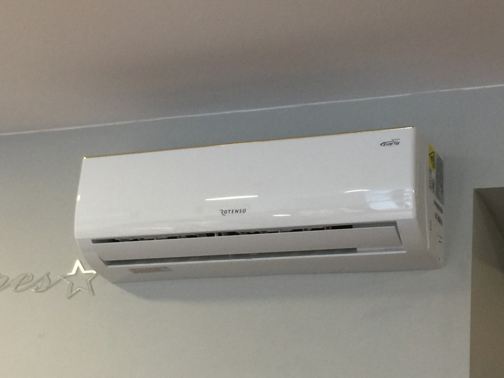 Biała jednostka wewnętrzna klimatyzacji Rotenso Inverter zamontowana na szarej ścianie, widoczne dekoracje ścienne w formie napisu i gwiazdy.