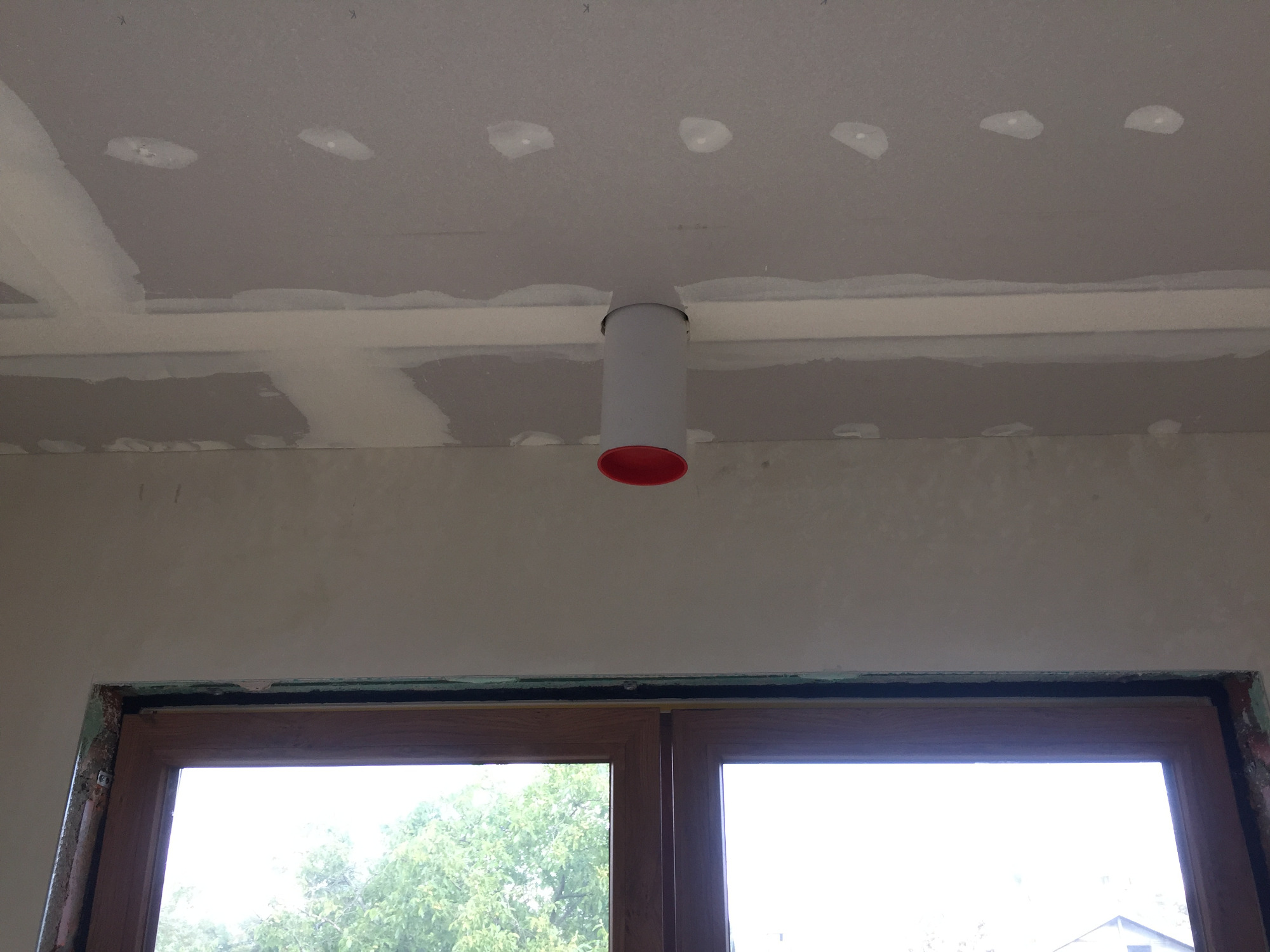 Szary kanał wentylacyjny z czerwoną końcówką zamontowany na suficie z płyt gipsowo-kartonowych, widok z dołu na okno.