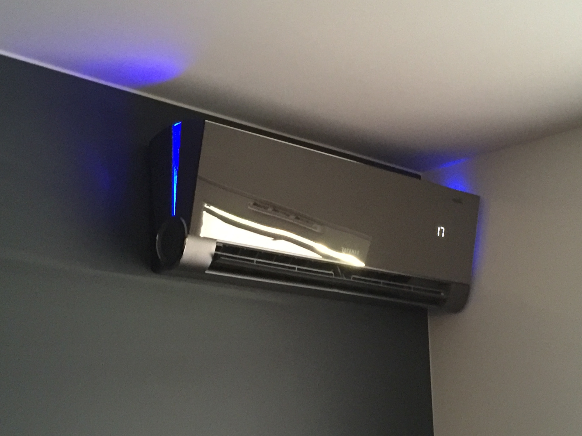 Szary klimatyzator ścienny z niebieskim podświetleniem LED na ścianie w dwukolorowym pokoju.