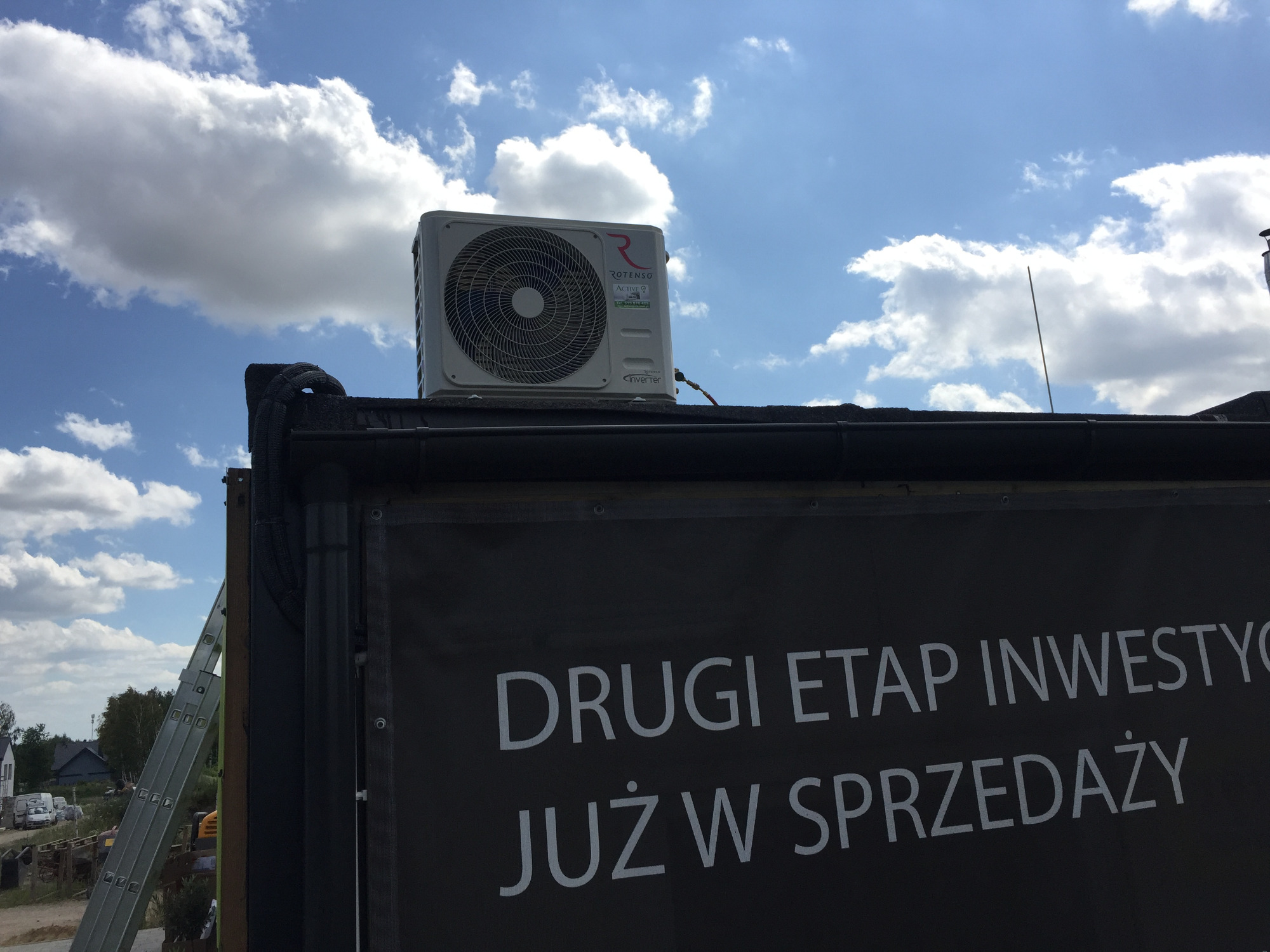Zewnętrzna jednostka klimatyzacji Rotenso Inverter zamontowana na dachu budynku, widoczny baner reklamowy 'Drugi etap inwestycji już w sprzedaży' i drabina oparta o ścianę, błękitne niebo w tle.