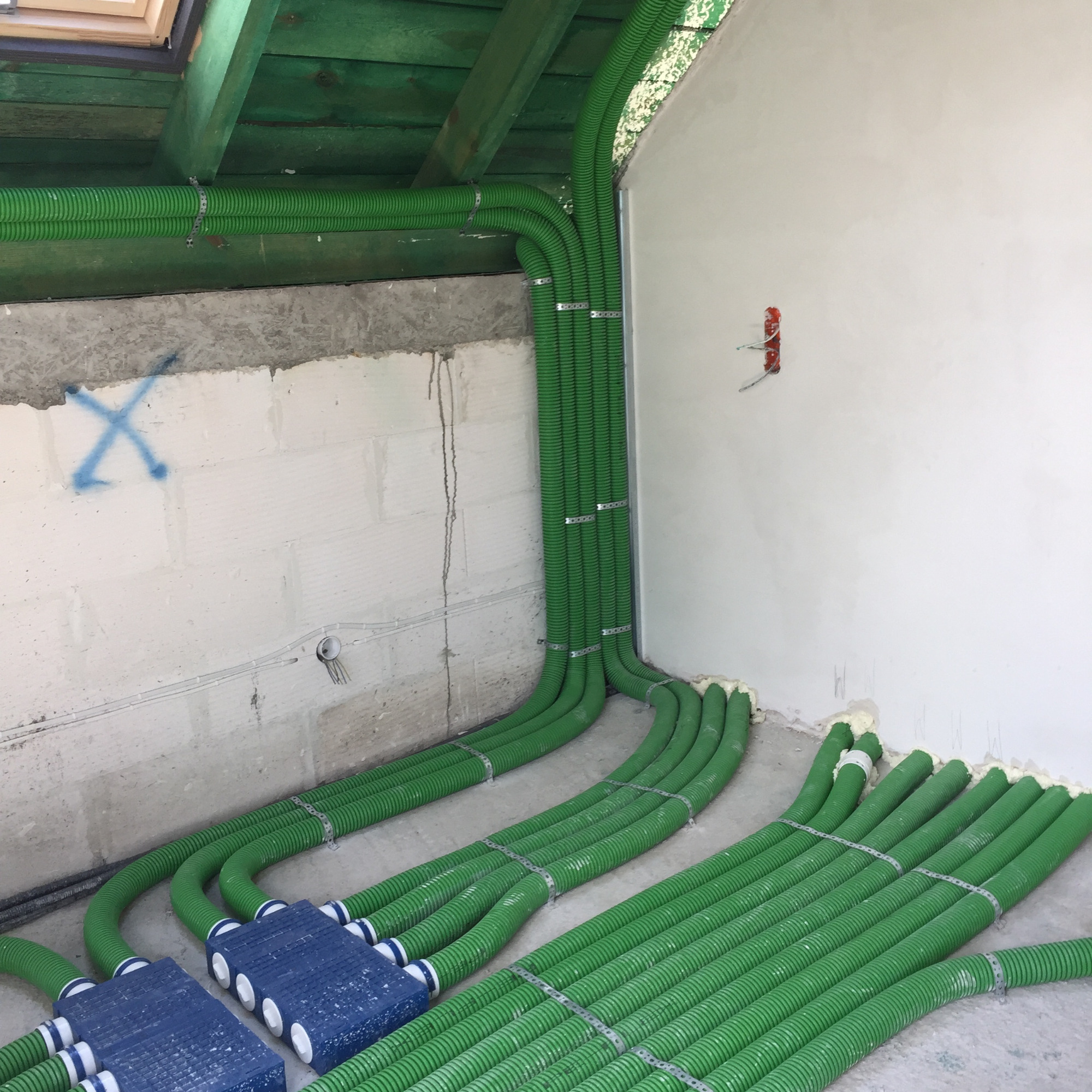 Zielone, karbowane rury wentylacyjne przymocowane metalowymi obejmami do surowych ścian i sufitu w trakcie instalacji w nowym budynku, widoczne niebieskie skrzynki rozdzielcze i pianka montażowa.