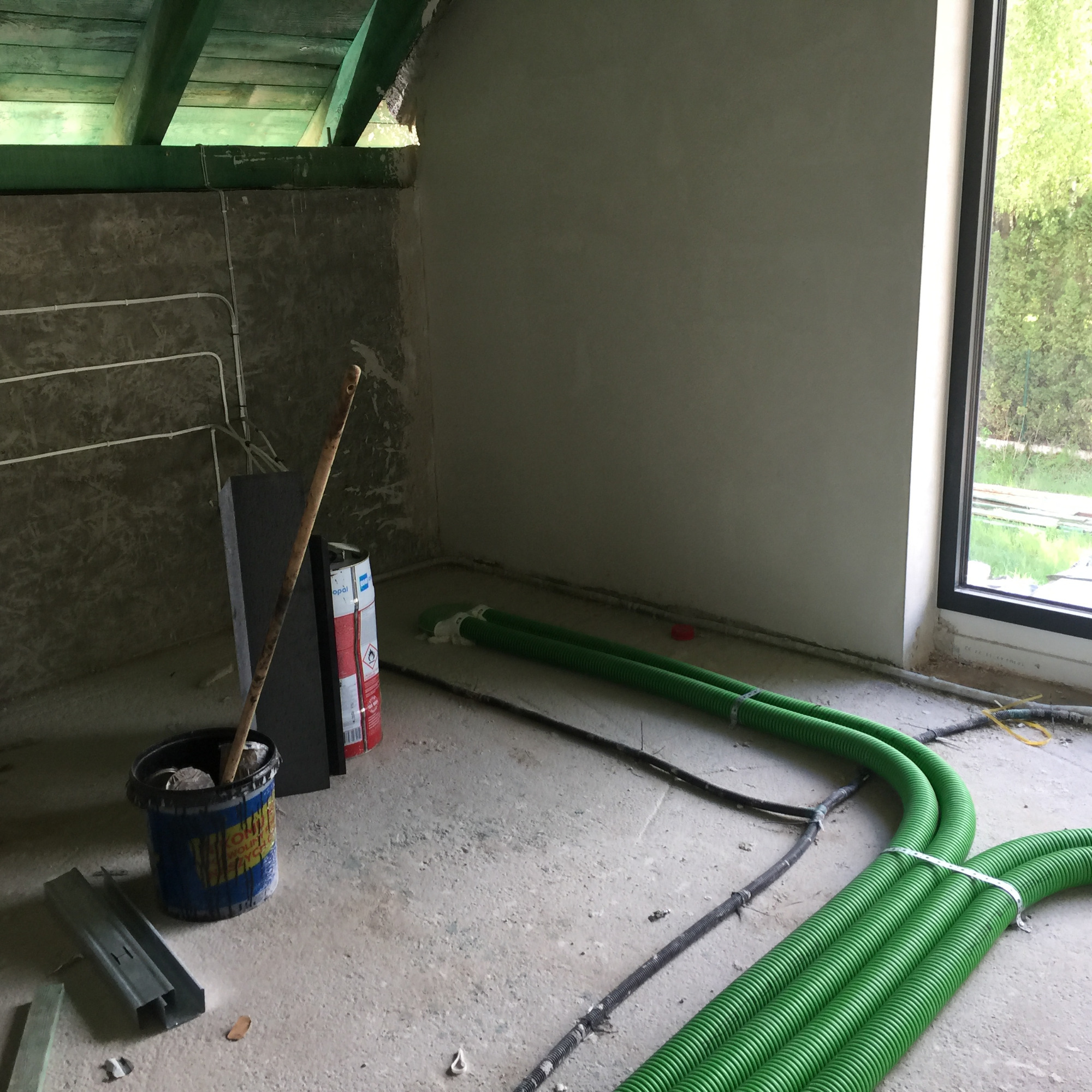 Wnętrze w trakcie prac wykończeniowych: widoczne zielone rury wentylacyjne, wiadro z narzędziami, puszka farby i fragment okna z widokiem na zieleń.
