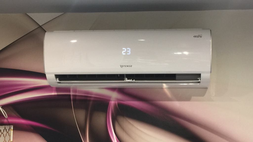 Biały klimatyzator Rotenso z wyświetlaczem temperatury 23 stopnie Celsjusza, zamontowany na ścianie z wzorem w odcieniach różu i fioletu.
