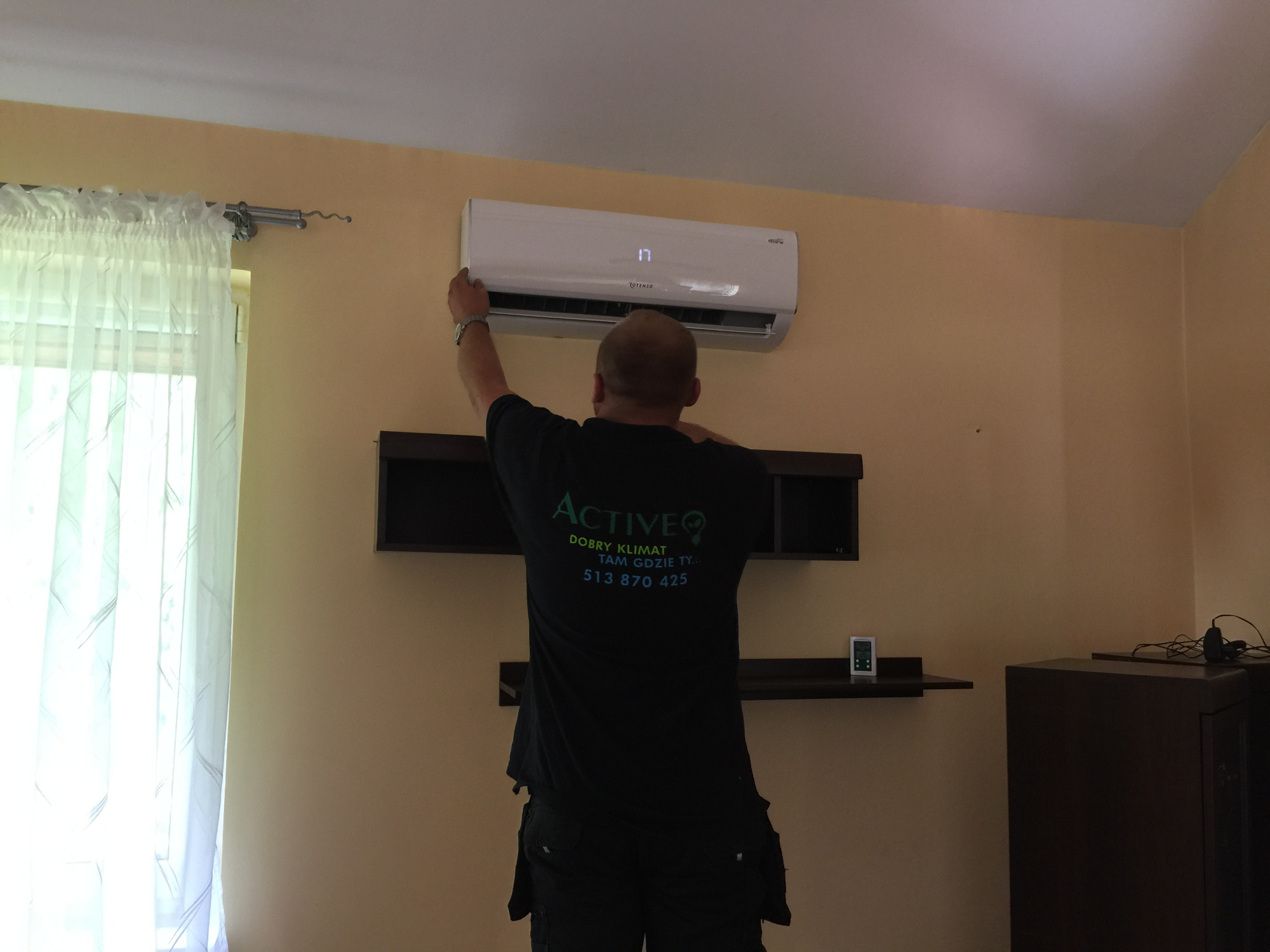 Instalator w czarnej koszulce z logo firmy montuje białą jednostkę wewnętrzną klimatyzacji na ścianie w pokoju z beżowymi ścianami.