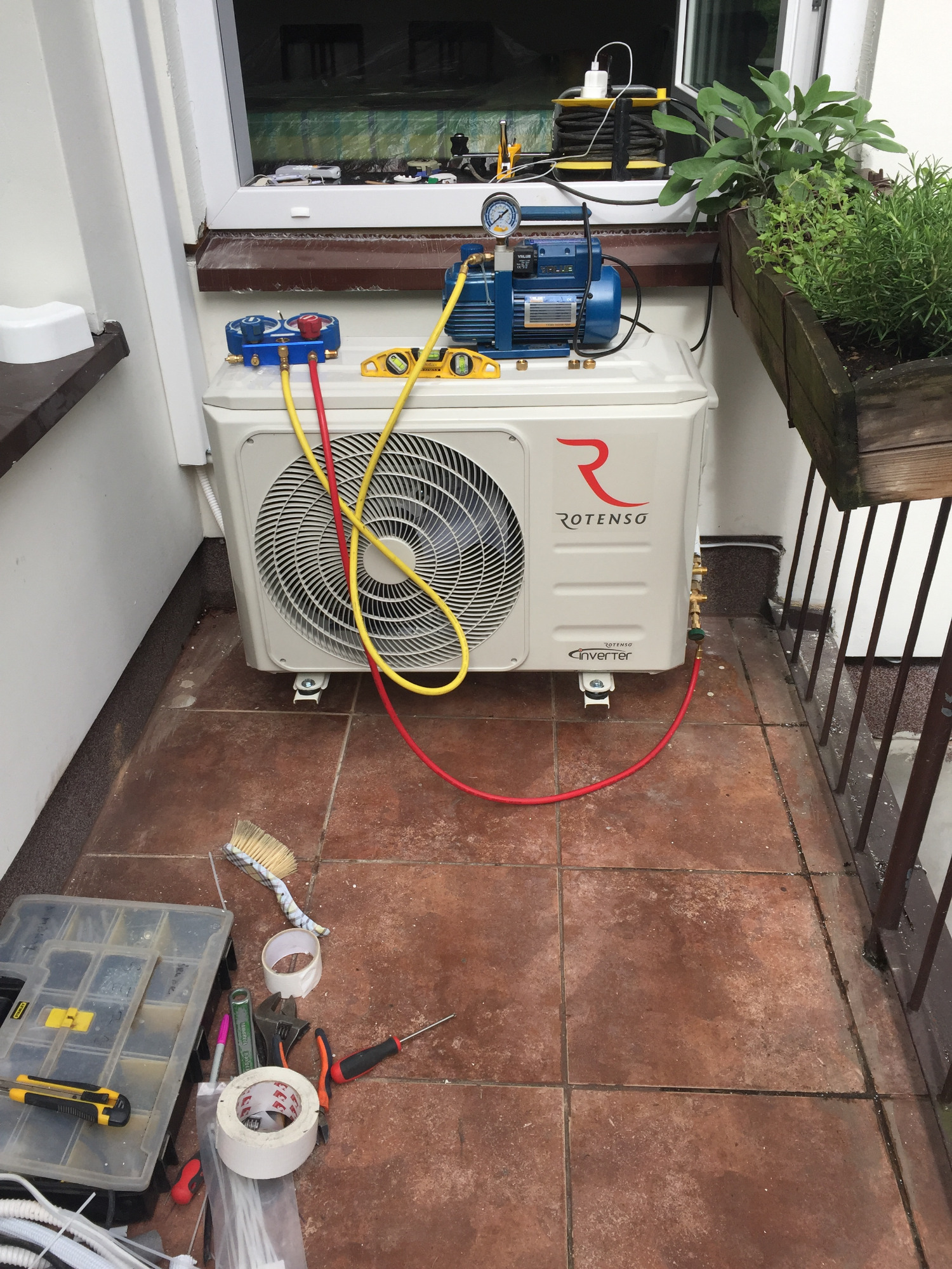 Biały klimatyzator Rotenso Inverter z podłączonymi przewodami serwisowymi i pompą próżniową na balkonie z brązowymi płytkami, obok skrzynka z narzędziami.