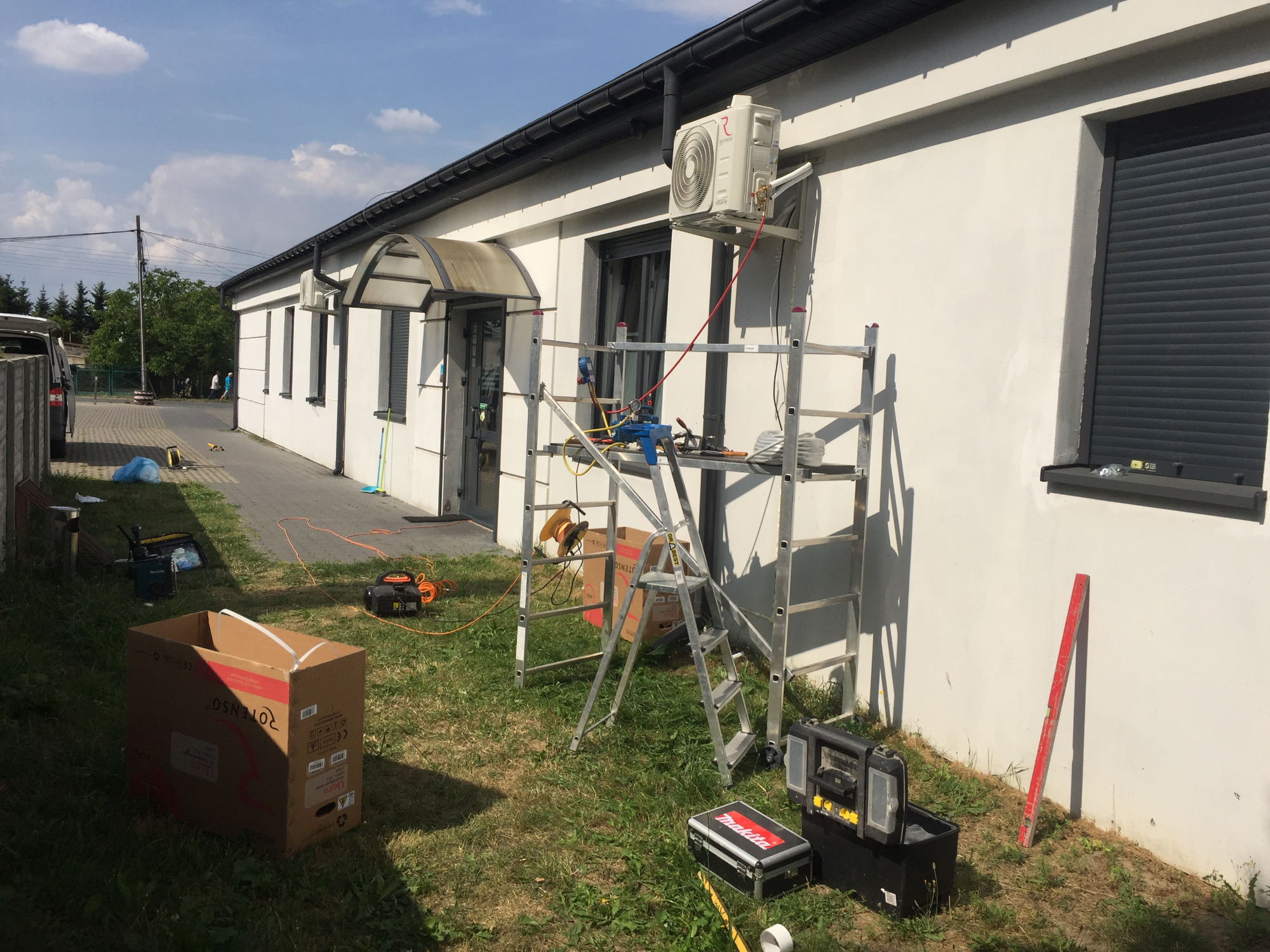 Biały budynek z zamontowaną jednostką zewnętrzną klimatyzatora, widoczne rusztowanie, drabina i narzędzia na trawie podczas instalacji.