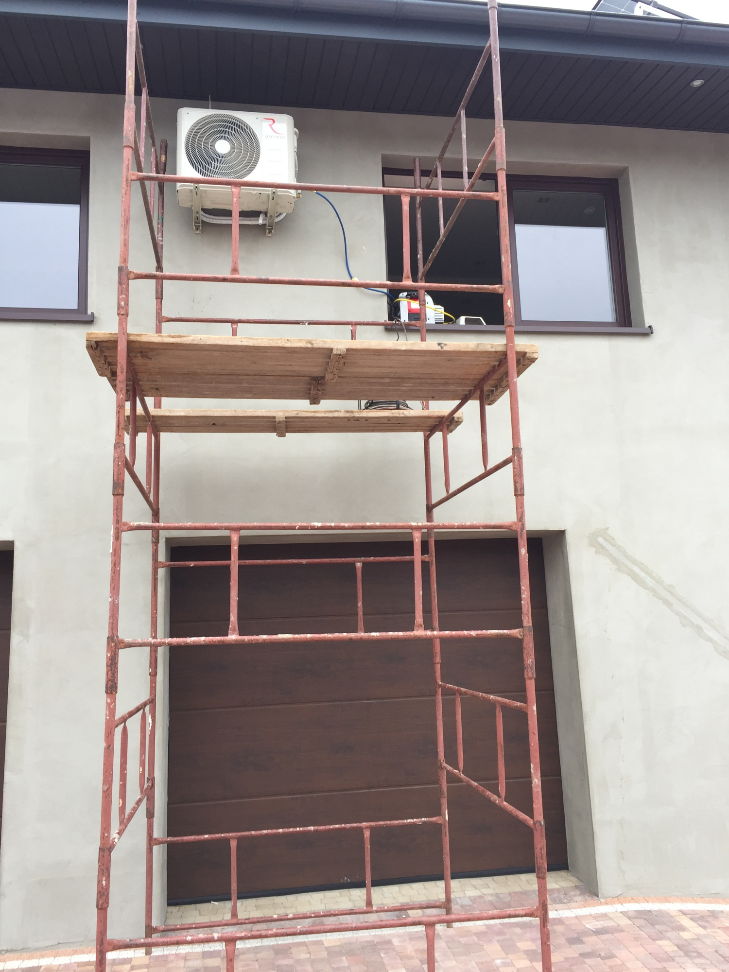 Zewnętrzna jednostka klimatyzatora zamontowana na elewacji budynku, widoczna z rusztowania, z narzędziami na platformie
