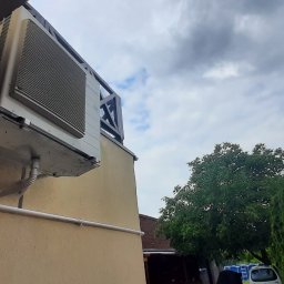 VESTIGO PC SPÓŁKA Z OGRANICZONĄ ODPOWIEDZIALNOŚCIĄ - Zewnętrzna jednostka pompy ciepła zamontowana na ścianie budynku w Woli Kopcowej, widok z dołu, instalacja z rurami odprowadzającymi skropliny.