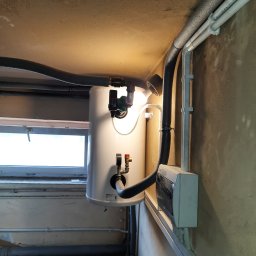 VESTIGO PC SPÓŁKA Z OGRANICZONĄ ODPOWIEDZIALNOŚCIĄ - Biały bojler elektryczny zamontowany na ścianie w piwnicy, widoczne rury, zawory, pompa obiegowa i instalacja elektryczna.