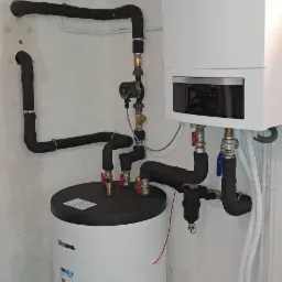 Biały piec gazowy Buderus i zasobnik ciepłej wody Kospel z czarnymi rurami instalacji grzewczej w narożniku piwnicy, widoczne zawory i pompy obiegowe.