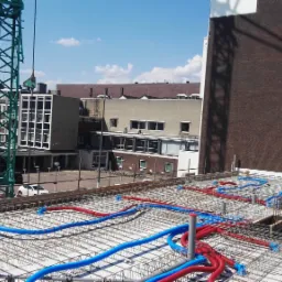 Instalacja rur wodnych i kanalizacyjnych, czerwonych i niebieskich, na zbrojeniu stropu budynku w trakcie budowy, z widocznym żurawiem budowlanym i zabudową miejską w tle.