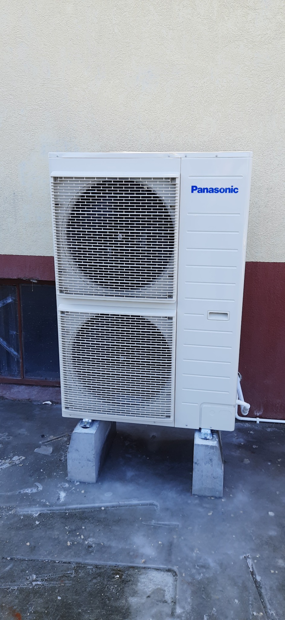 Zewnętrzna jednostka klimatyzacji Panasonic, zamontowana na betonowych podstawach przy ścianie budynku z widocznym oknem piwnicznym.