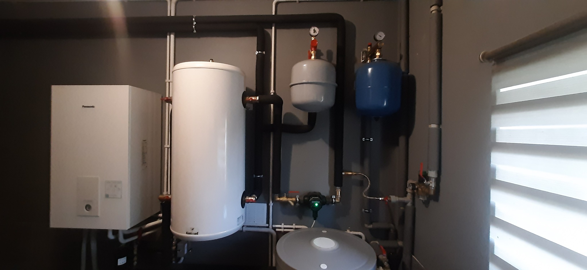 Zmontowana instalacja grzewcza: biały piec gazowy Panasonic, pionowy biały zbiornik, szary i niebieski zbiornik wyrównawczy, czarne rury izolowane, szare ściany, widoczne zawory i manometry.