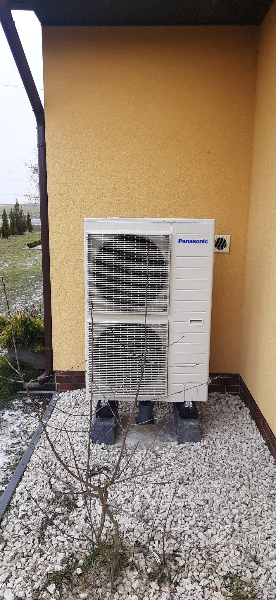 Zewnętrzna jednostka klimatyzacyjna Panasonic zamontowana na betonowych podstawach, na tle żółtej elewacji budynku i kamiennego podłoża, z widocznym fragmentem rynny i młodym drzewkiem na pierwszym...