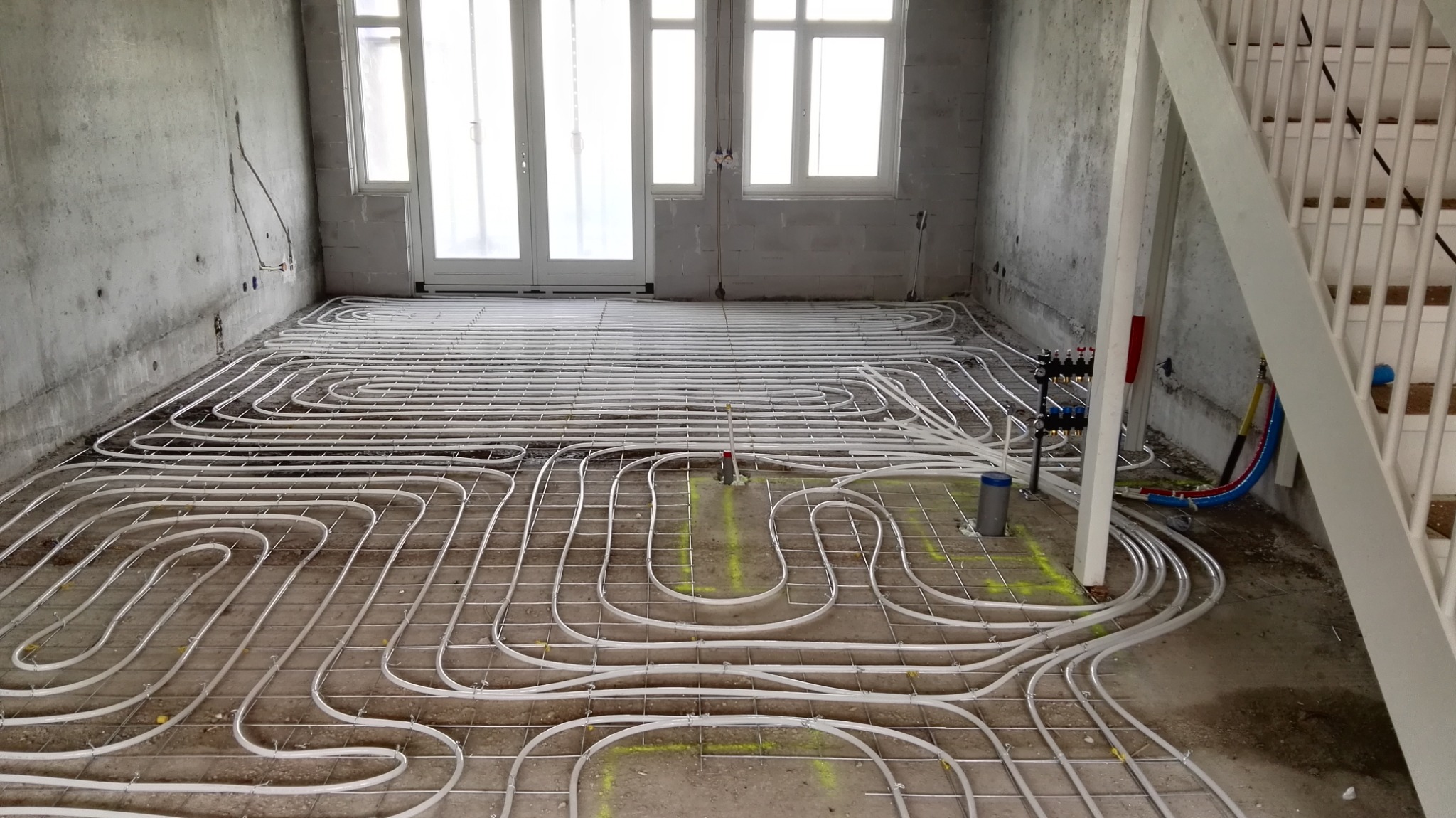 Rozłożone rury ogrzewania podłogowego na siatce w surowym wnętrzu, widoczne przyłącza i rozdzielacz, przygotowane do zalania wylewką.