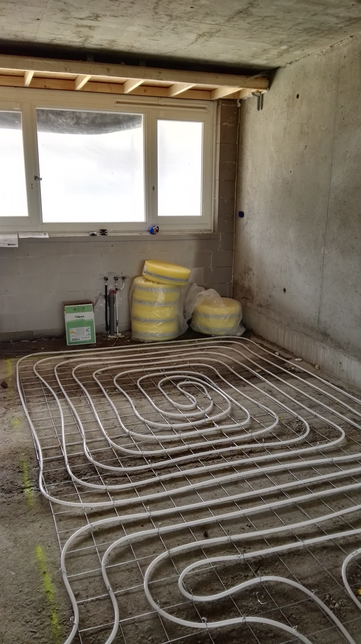 Instalacja ogrzewania podłogowego w surowym stanie, widoczne rury grzewcze ułożone na metalowej siatce, izolacja termiczna w żółtej folii obok okna, ściany z surowego betonu.