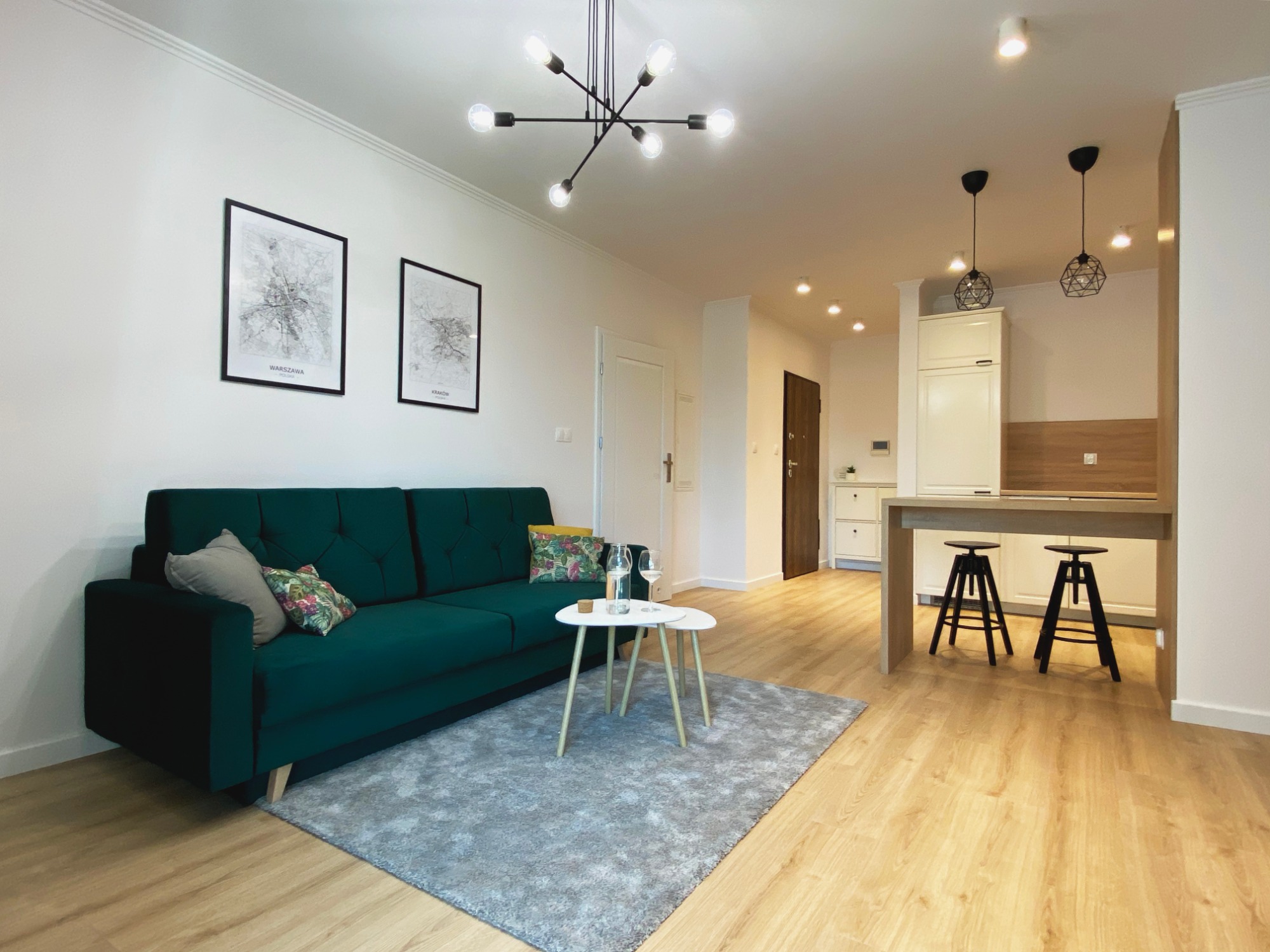 Nowoczesne wnętrze: Szmaragdowa sofa z poduszkami, stolik kawowy na tle szarego dywanu, drewniana podłoga, plakaty z mapami Warszawy i Krakowa, industrialna lampa sufitowa, aneks kuchenny z hokerami.