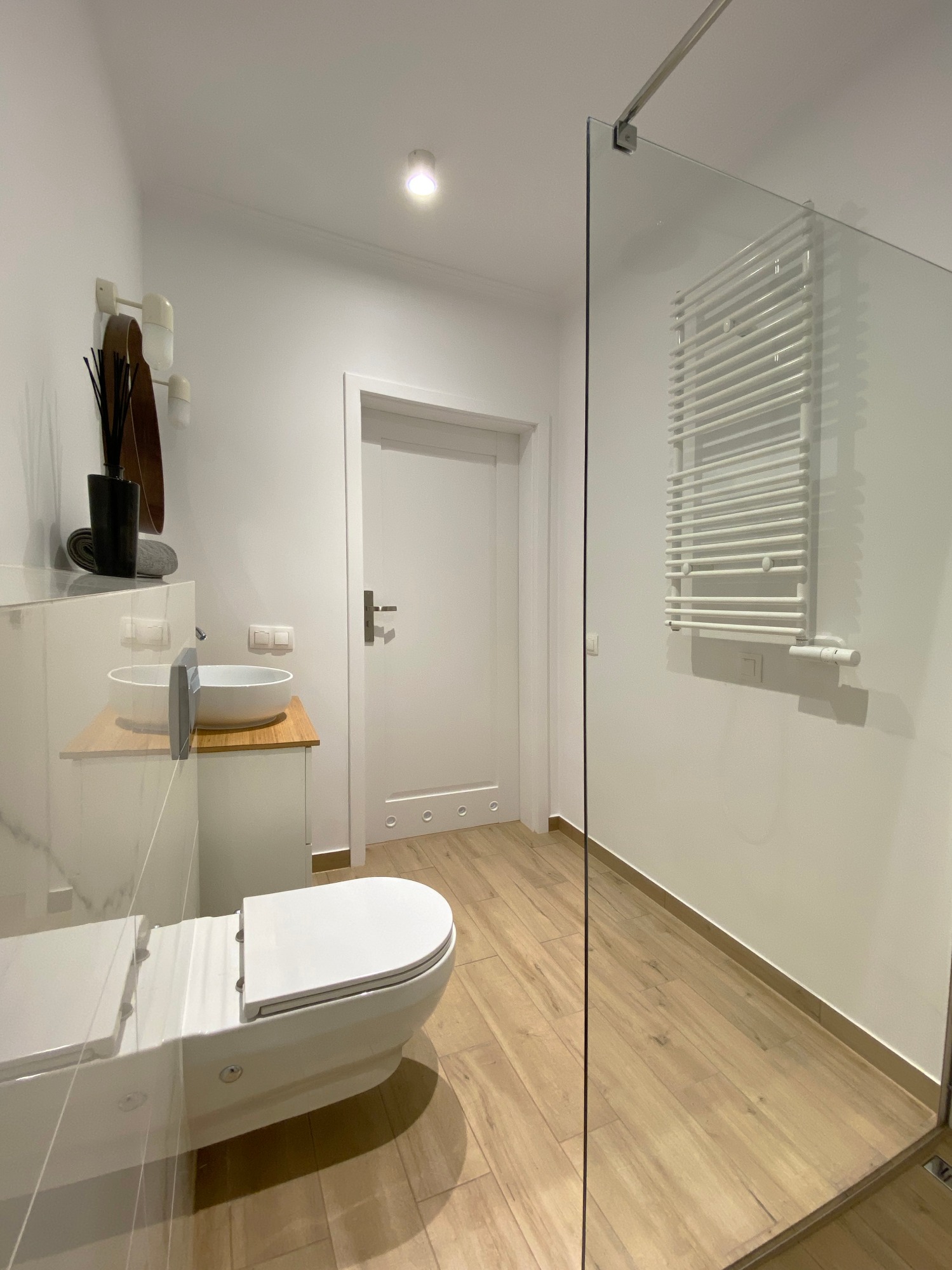Nowoczesna łazienka w jasnych barwach z drewnianą podłogą i szklaną kabiną prysznicową, widoczna umywalka nablatowa i wisząca toaleta.