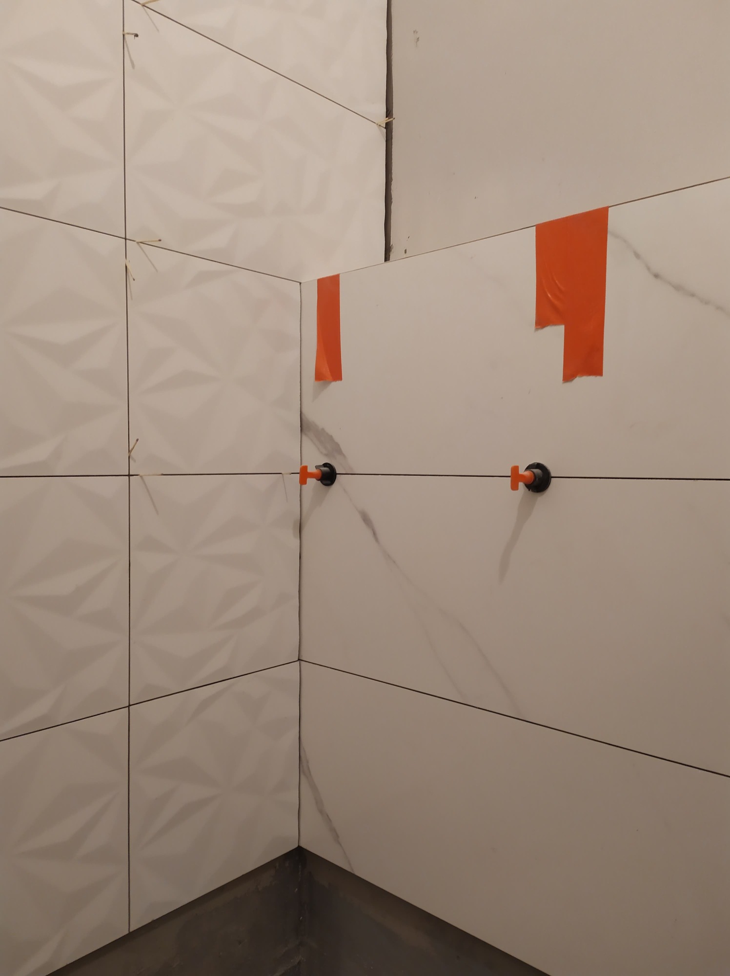 Fragment ściany w trakcie układania białych, trójwymiarowych płytek w łazience, widoczne pomarańczowe paski taśmy i czarne kliny poziomujące płytki.