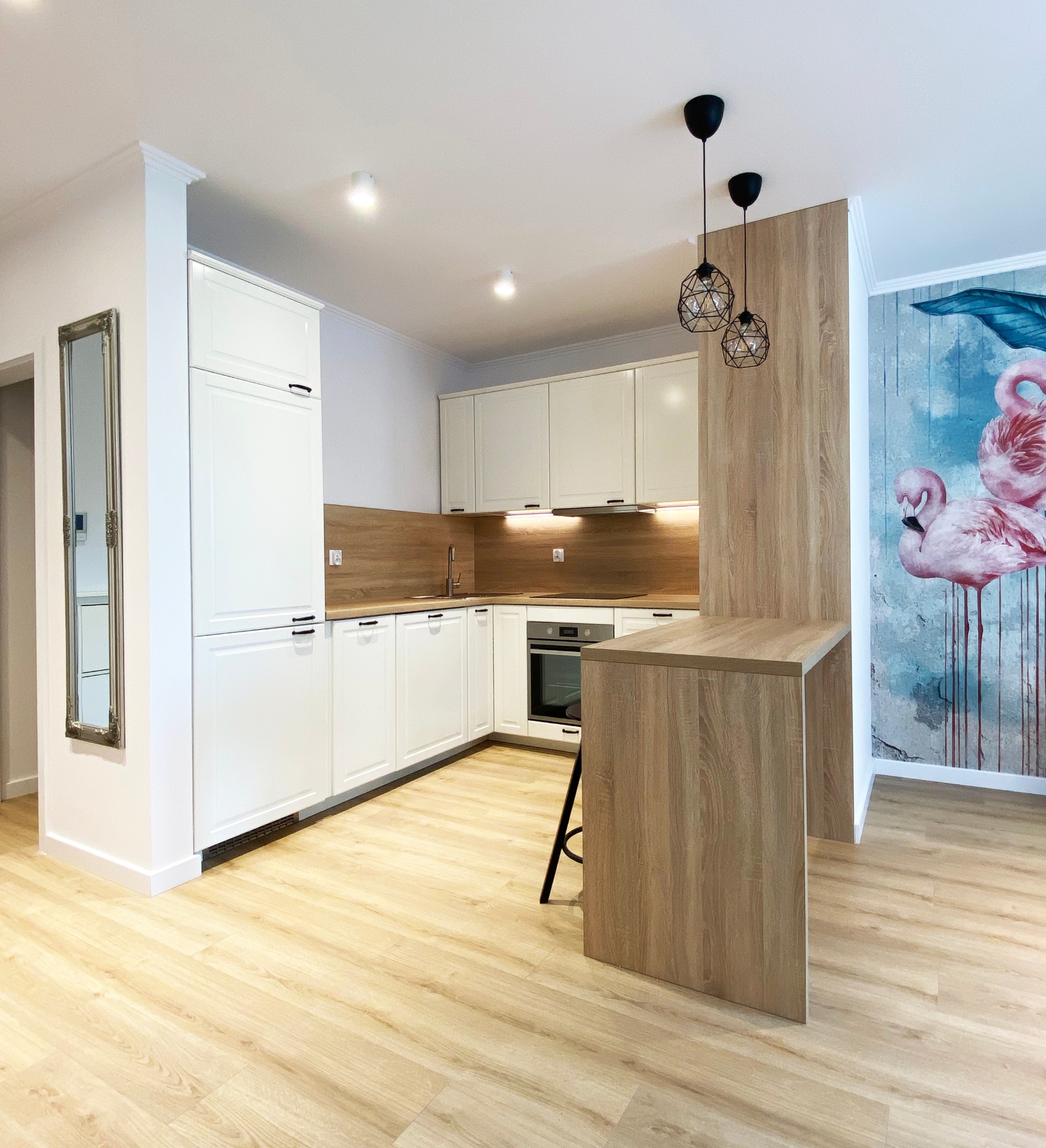 Jasna kuchnia w stylu nowoczesnym z białymi szafkami, drewnianym blatem i wyspą; na ścianie tapeta z motywem flamingów, a na podłodze panele imitujące drewno.