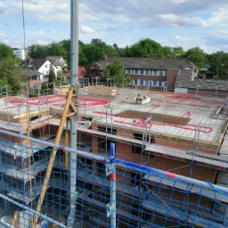 Wysoki kąt widzenia na plac budowy z widocznym zbrojeniem na stropie, czerwone rury instalacyjne, rusztowania Layher i ochronna siatka w kolorze niebieskim.