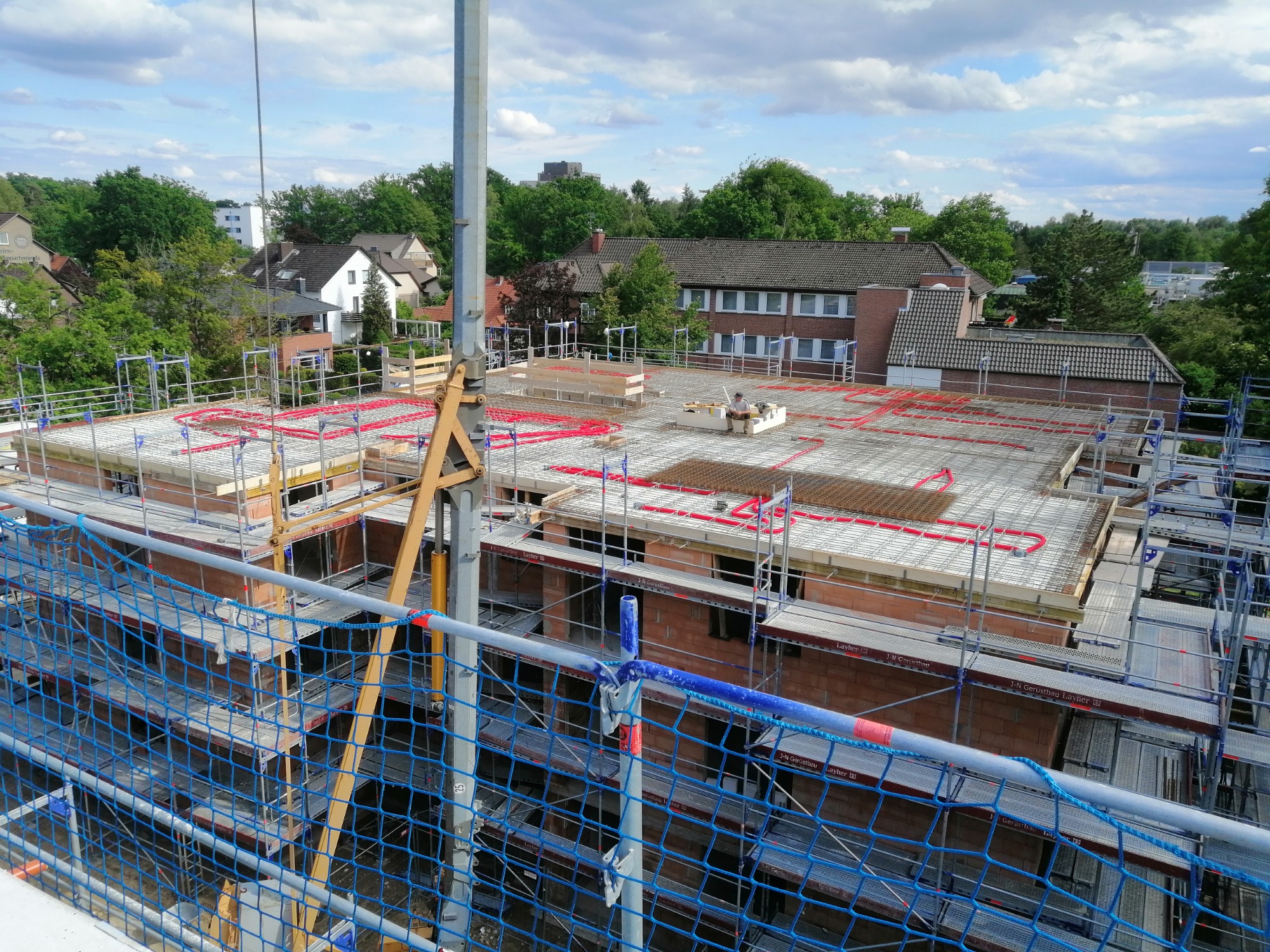 Wysoki kąt widzenia na plac budowy z widocznym zbrojeniem na stropie, czerwone rury instalacyjne, rusztowania Layher i ochronna siatka w kolorze niebieskim.