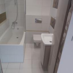 Firma remontowo budowlana Leszek Grabowski - Ukończona łazienka z białą wanną, umywalką z szafką i toaletą, wykończona biało-beżowymi płytkami, widoczny fragment surowej ściany przy drzwiach.