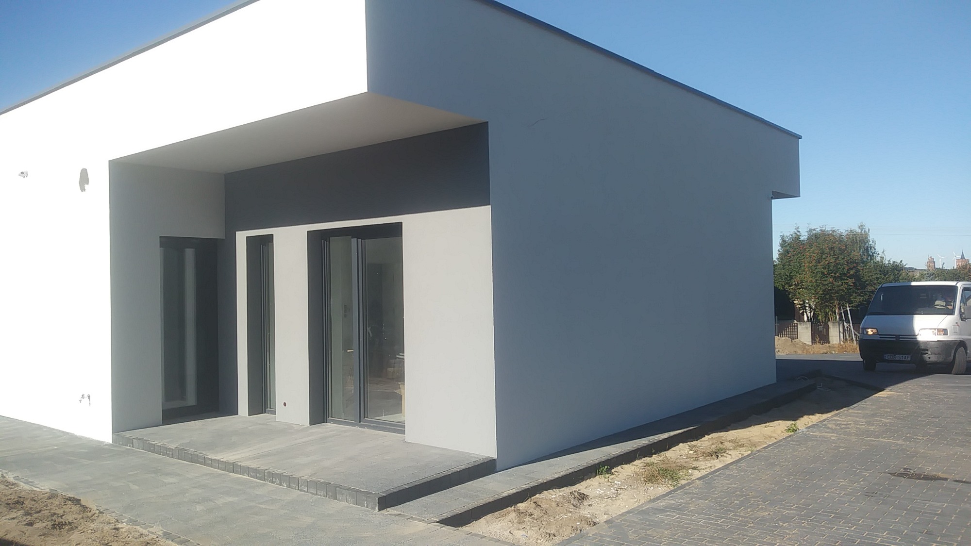 Nowoczesny dom jednorodzinny z szarą elewacją i ciemnymi ramami okiennymi, widoczny z zewnątrz, z białym fragmentem fasady po lewej stronie, na pierwszym planie kostka brukowa, a w tle biały...