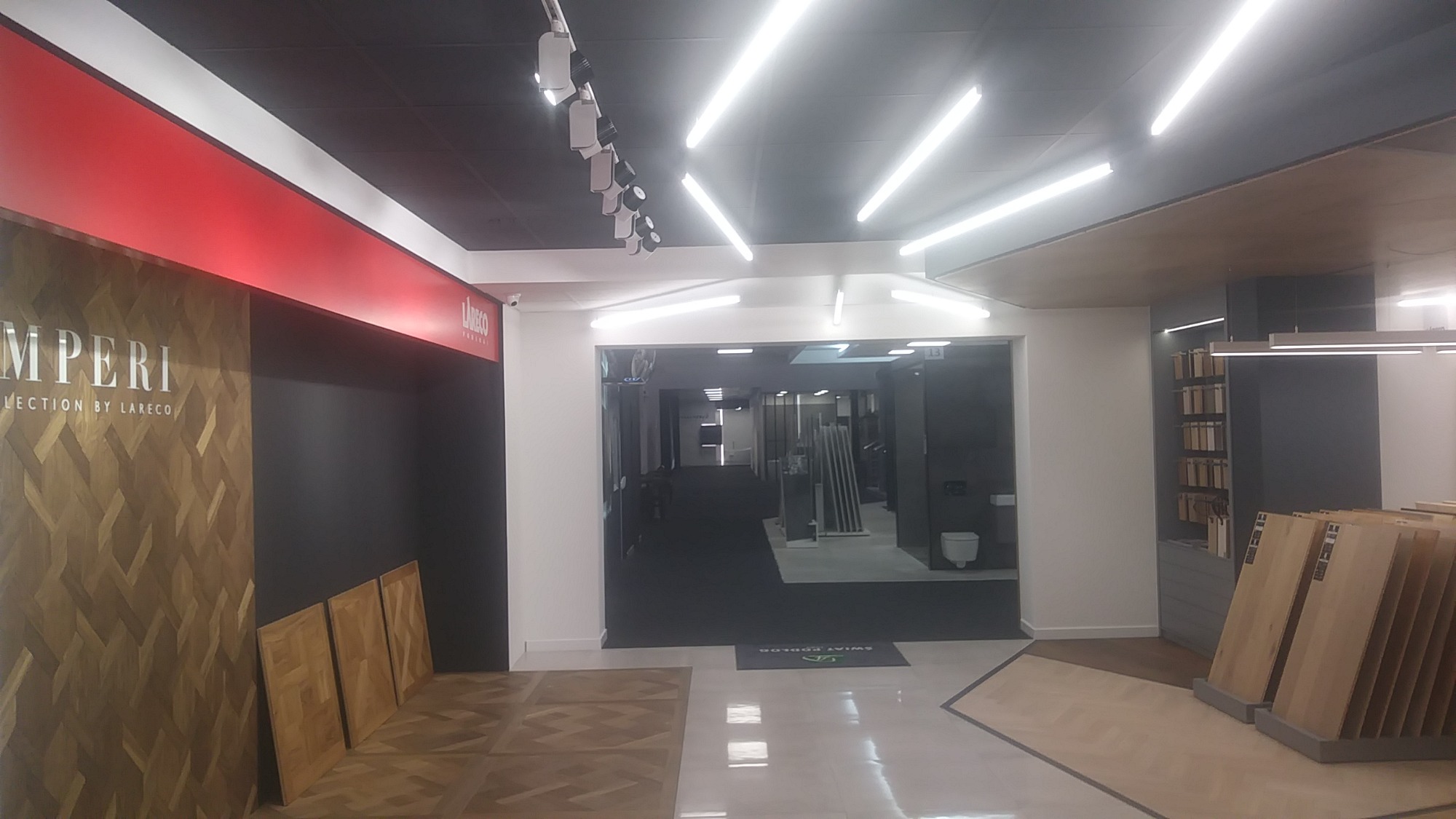 Wnętrze salonu z ekspozycją paneli podłogowych z drewna w różnych wzorach i kolorach, oświetlenie sufitowe LED, widoczne logo 'Imperio Collection by Lareco'.