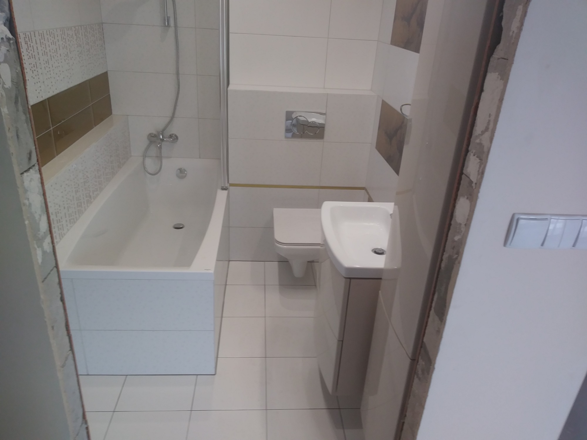 Ukończona łazienka z białą wanną, umywalką z szafką i toaletą, wykończona biało-beżowymi płytkami, widoczny fragment surowej ściany przy drzwiach.