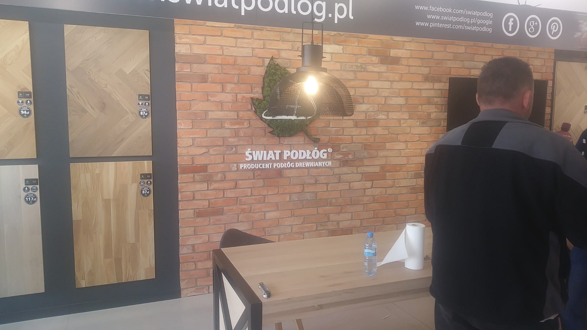 Ekspozycja paneli podłogowych w salonie Świat Podłóg, z widocznymi próbkami na ścianie, ceglanym tłem z logo firmy i lampą, oraz fragmentem stołu z butelką wody i rolką papieru.