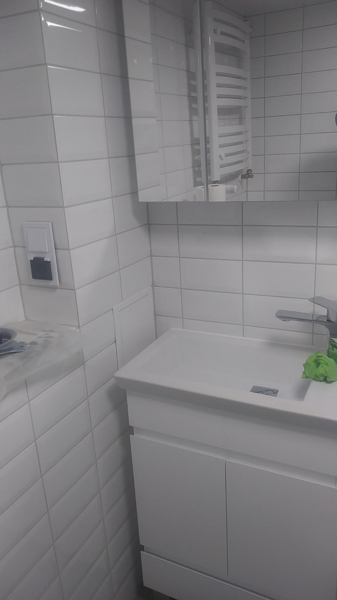 Biała umywalka z szafką w nowoczesnej łazience, widoczny grzejnik drabinkowy, białe płytki na ścianach i gniazdko elektryczne z szarą wkładką.