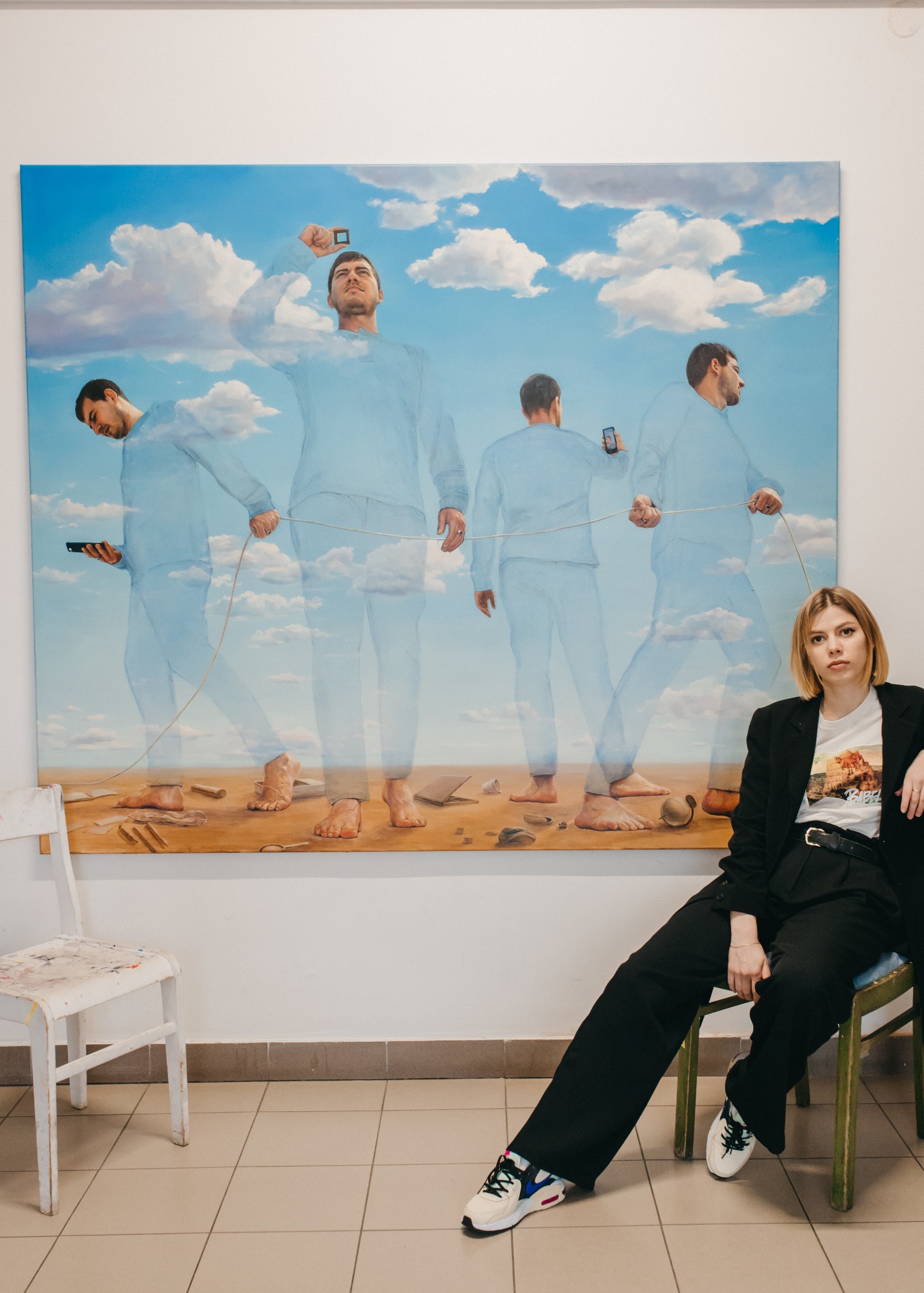 Obraz olejny 160/200cm przedstawiający mężczyzn w błękitnych strojach na tle nieba i piasku, trzymających linę i smartfony, z modelką siedzącą na krześle na pierwszym planie.
