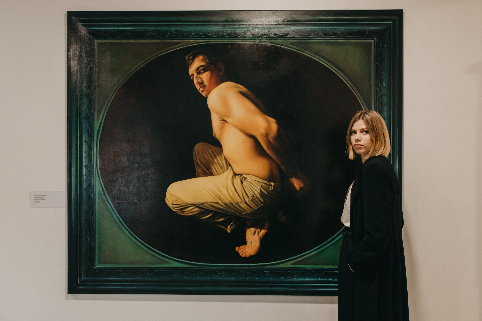 Duży obraz olejny przedstawiający nagiego mężczyznę w pozycji skulonej, w sepiowych spodniach, w ozdobnej, ciemnozielonej ramie; obok stoi blondynka w czarnym płaszczu.
