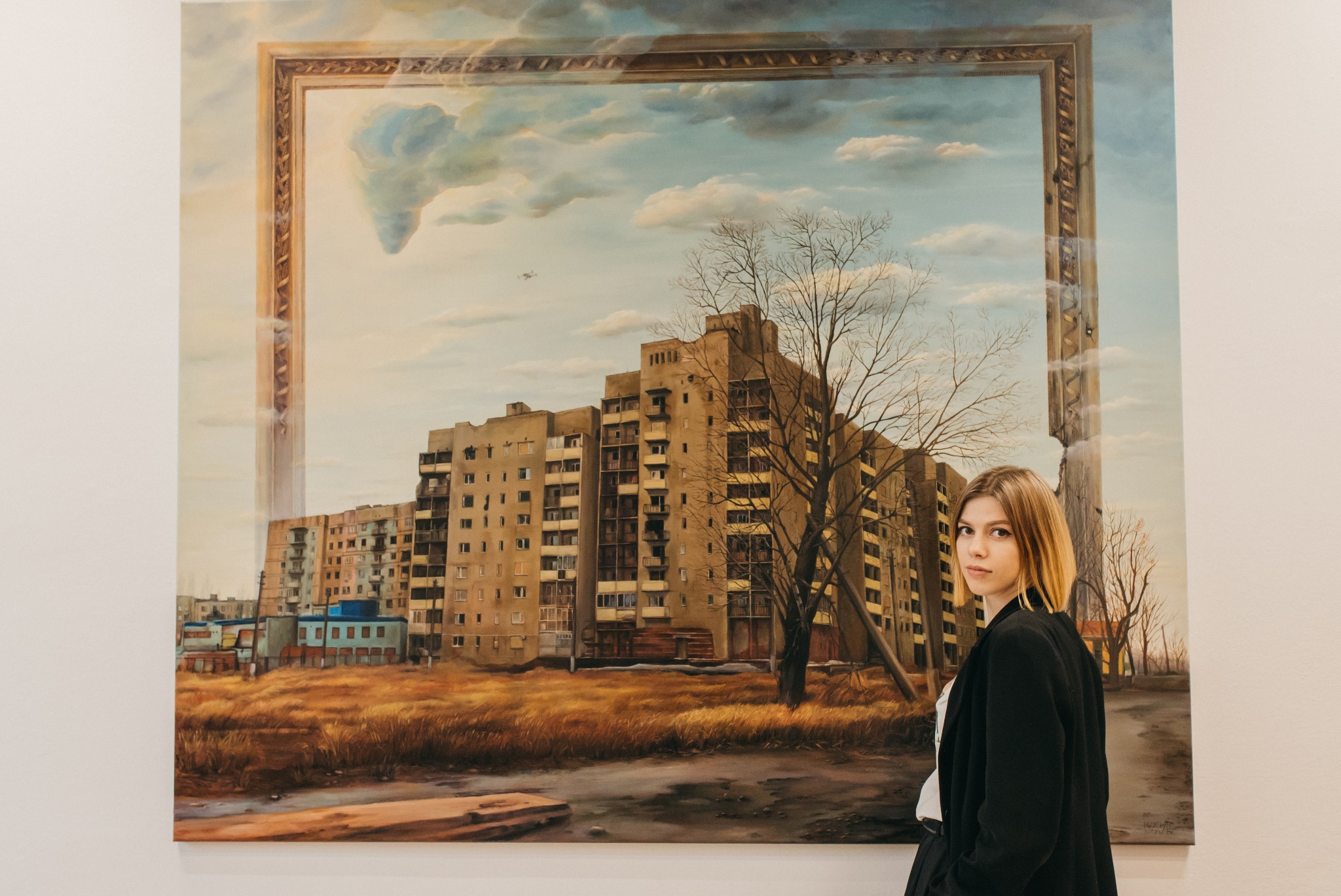 Obraz olejny na płótnie przedstawiający blokowisko w ramie, z młodą kobietą w czarnej marynarce stojącą obok. W tle błękitne niebo z chmurami i sylwetka samolotu.
