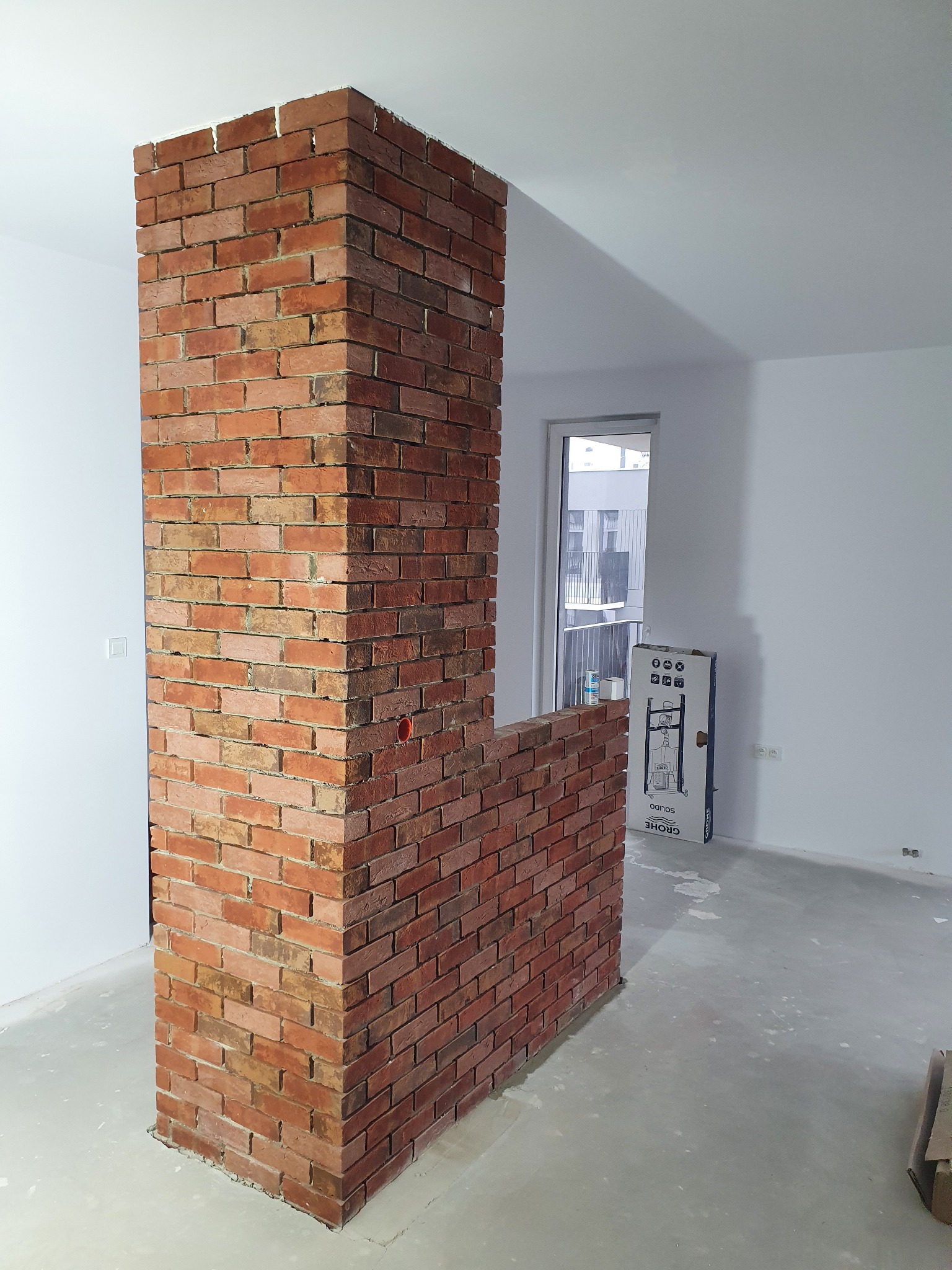 Niska ścianka działowa z cegły klinkierowej w surowym stanie, widoczne niedociągnięcia w spoinowaniu, białe ściany i surowa podłoga w tle, karton z armaturą sanitarną przy ścianie.