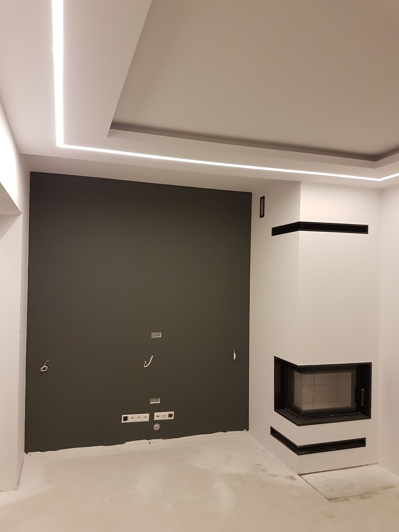 Nowoczesne wnętrze w trakcie wykańczania: szara ściana z gniazdkami, biały kominek z czarnymi akcentami i oświetlenie LED w suficie.