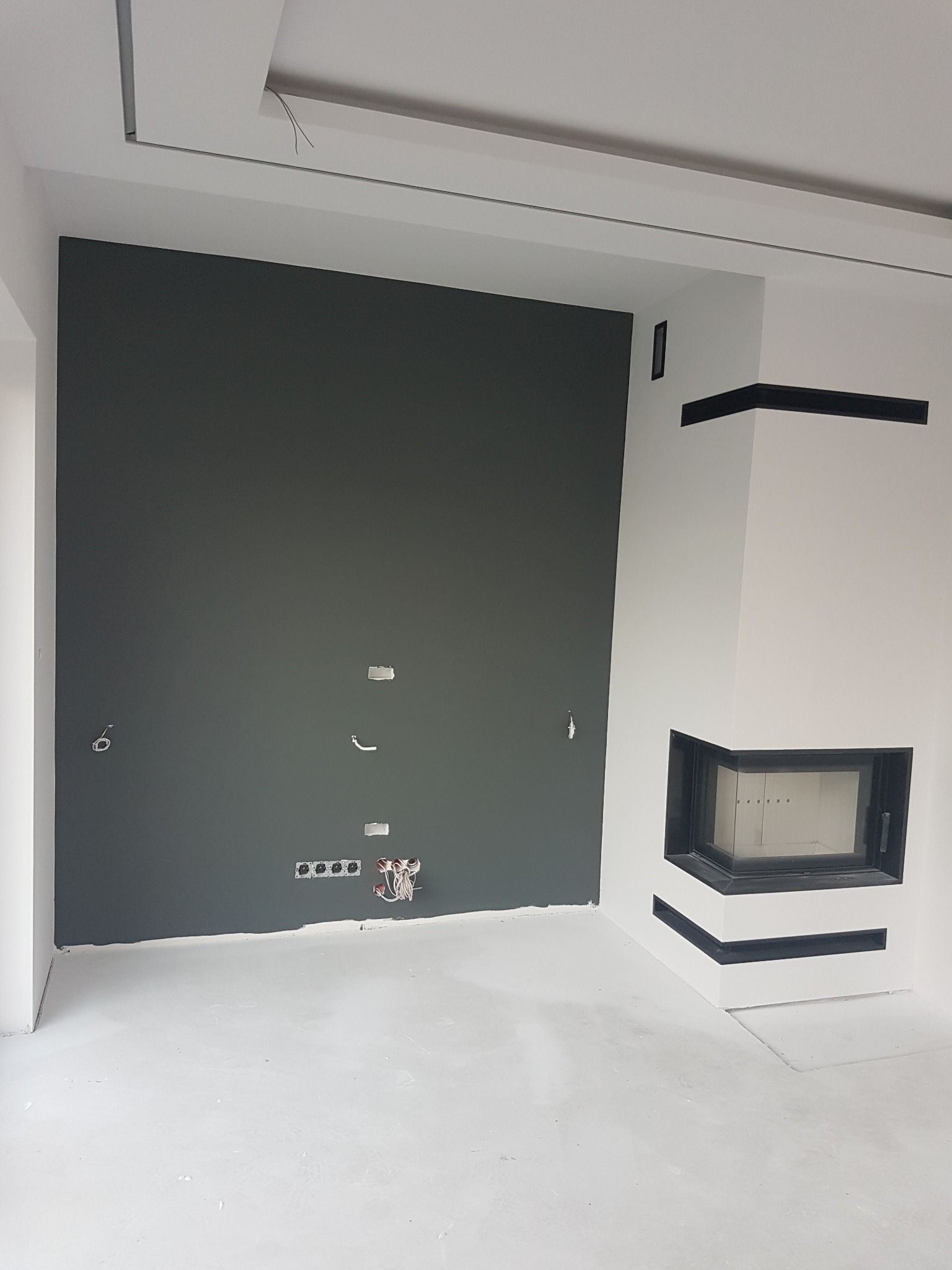 Szara ściana z przygotowanymi punktami elektrycznymi i gniazdkami, obok biały kominek z czarnymi akcentami w surowym wnętrzu w trakcie wykończenia.