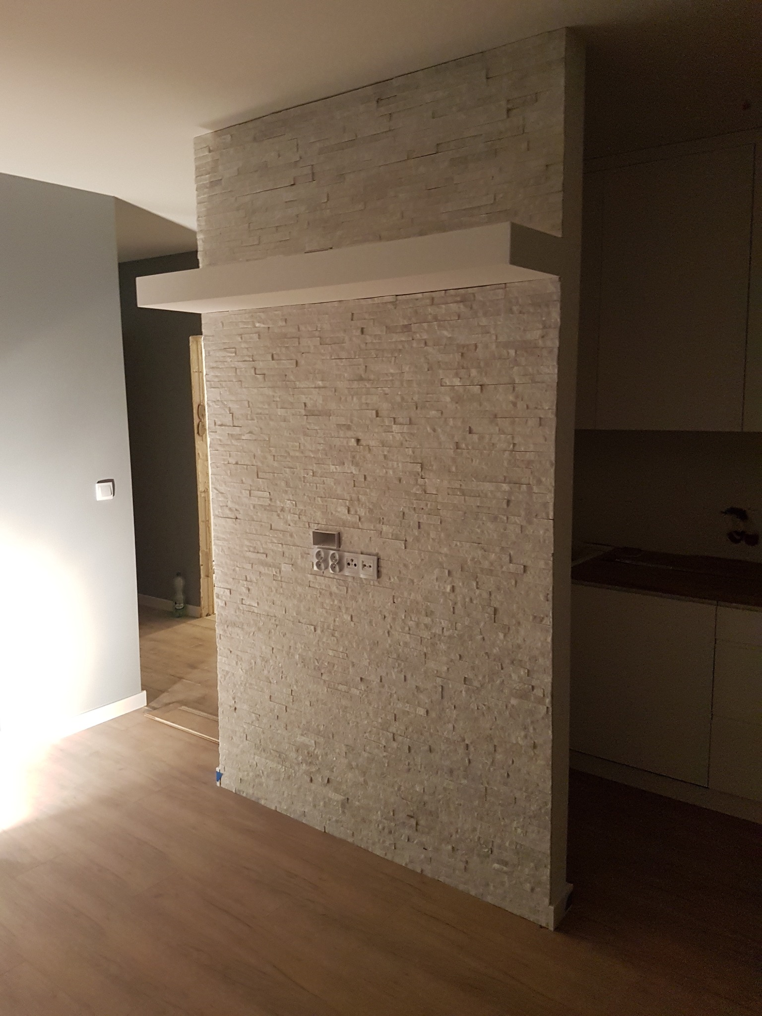 Ścianka działowa z dekoracyjnej cegły z półką, gniazdka elektryczne i włączniki zamontowane na cegle, widoczna podłoga z paneli i fragment kuchni w tle.