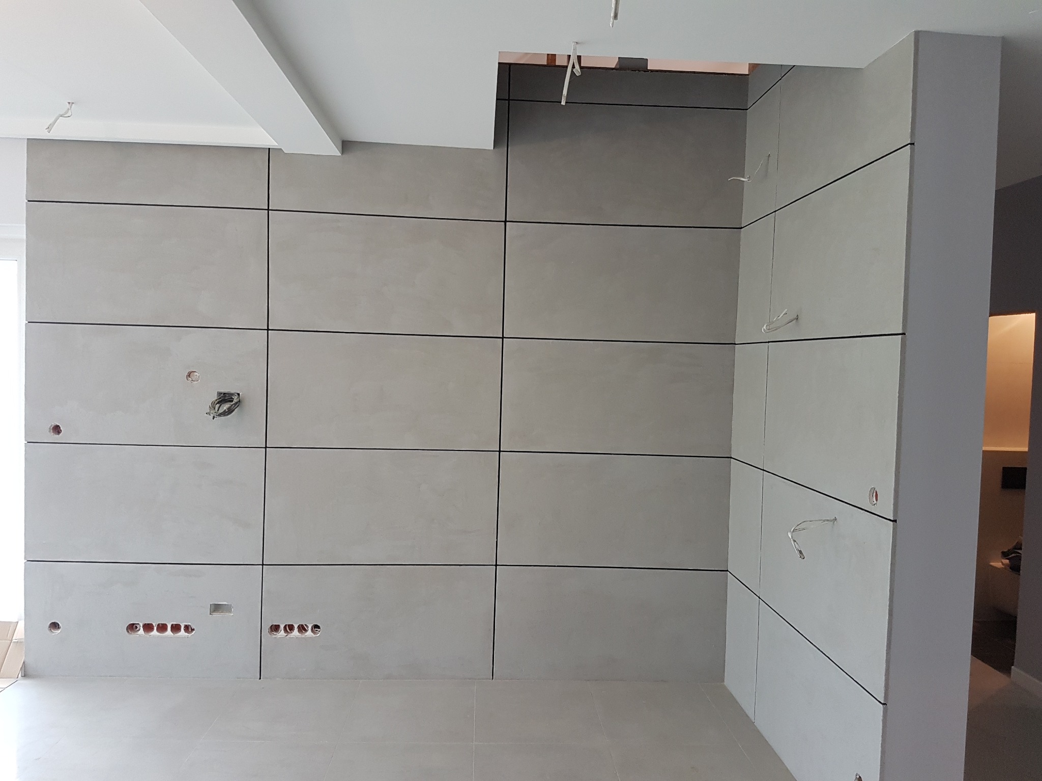 Ściana wykończona panelami w odcieniach szarości z widocznymi otworami na gniazdka elektryczne i wyprowadzeniami kabli, surowy stan wykończenia wnętrza.