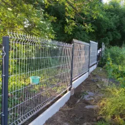 Montaż siatki ogrodzeniowej z podmurówką betonową, widoczny zielony pojemnik i częściowo osoba w tle, świeżo wykopana ziemia.