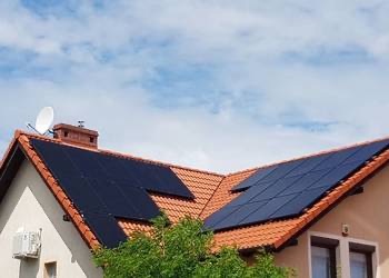 Ciemne panele na dachu domu jednorodzinnego z czerwoną dachówką, widok z dołu na błękitne niebo z chmurami.