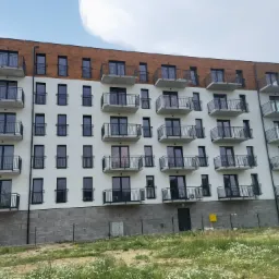 Nowoczesny budynek wielorodzinny z białymi ścianami, drewnianą elewacją na górze i czarnymi balkonami, widziany z poziomu gruntu, otoczony łąką z białymi kwiatami.