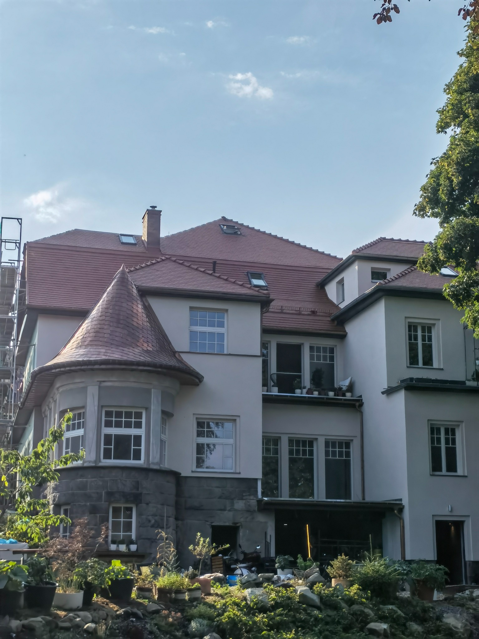 Wielopiętrowy budynek z jasną elewacją, czerwoną dachówką i wieżyczką z łupka, widoczne rusztowania z boku, w tle błękitne niebo i zieleń drzew.